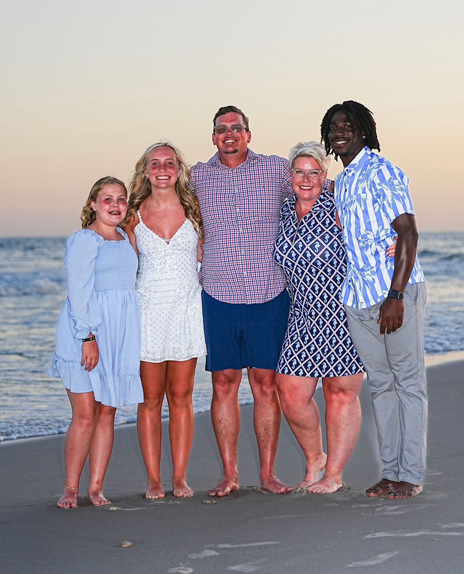 Five people smiling together on a beach at sunset, posing for a family portrait near the ocean shoreline.