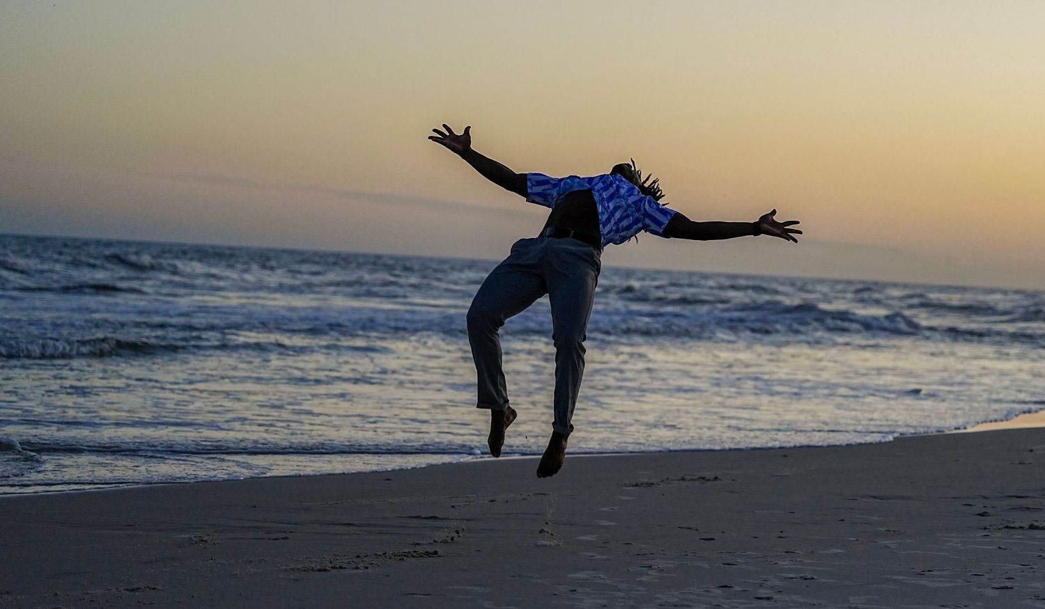Person jumping joyfully on a beach at sunset with arms outstretched, waves and colorful sky in the background.
