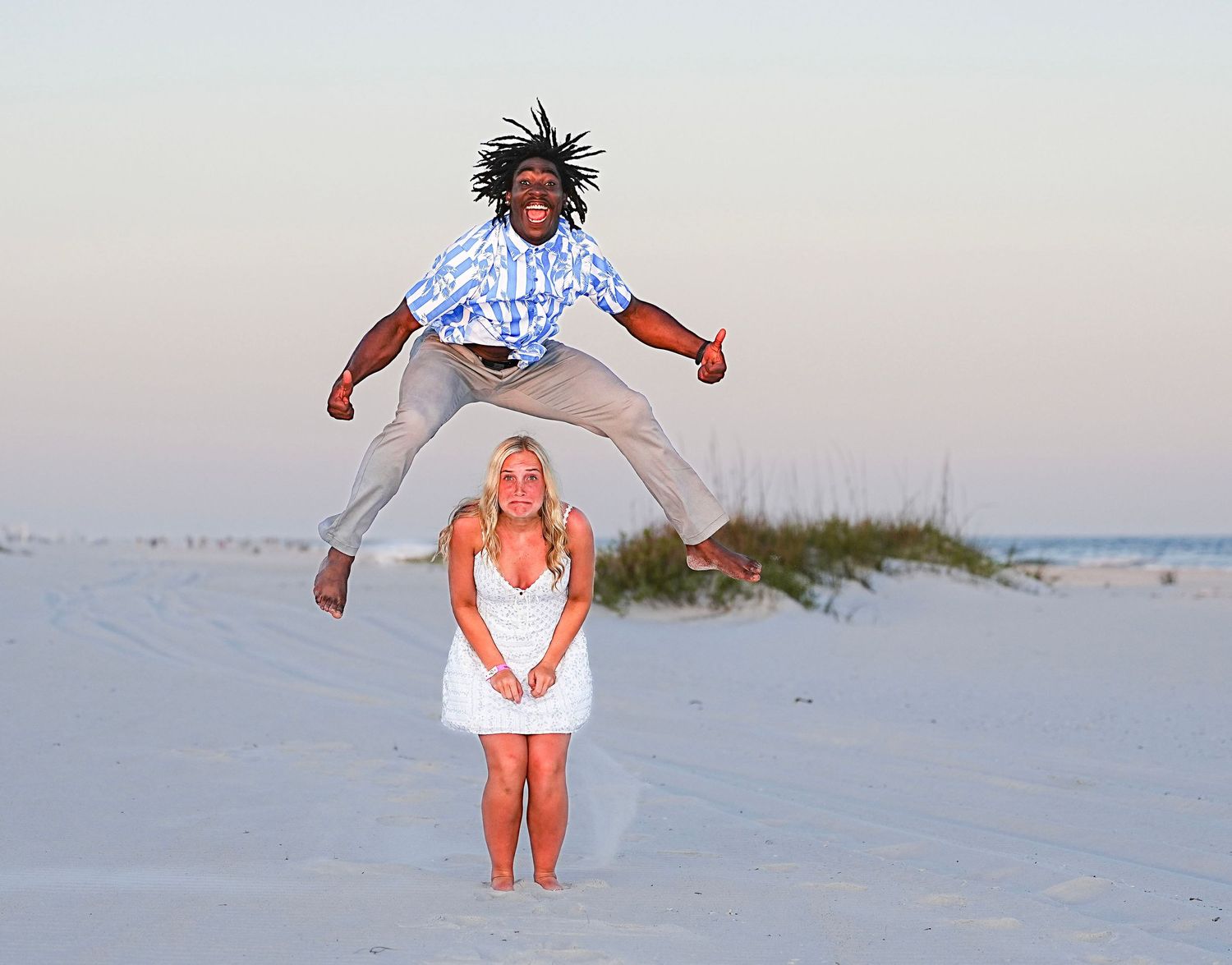 Man jumping high above woman in white dress on sandy beach at sunset.