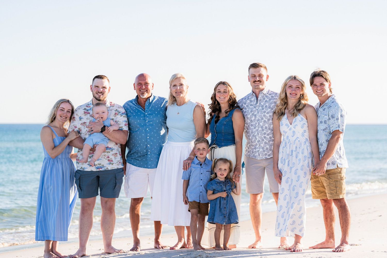 Family of nine poses smiling on a sunny beach, dressed in coordinating blue and white outfits, with calm ocean waves behind them.