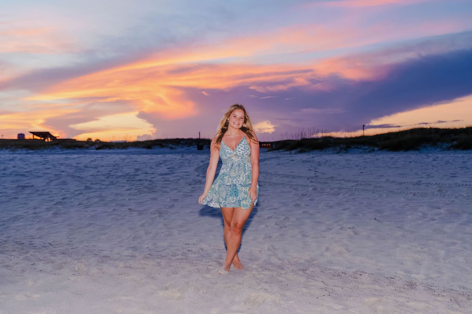 Blonde woman in floral romper stands barefoot on white sand beach at stunning pink and purple sunset.