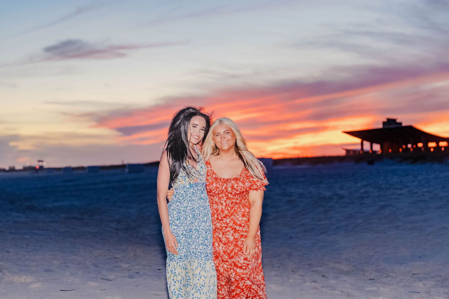 Two women in floral dresses smile together on a sandy beach during a vibrant pink and orange sunset.