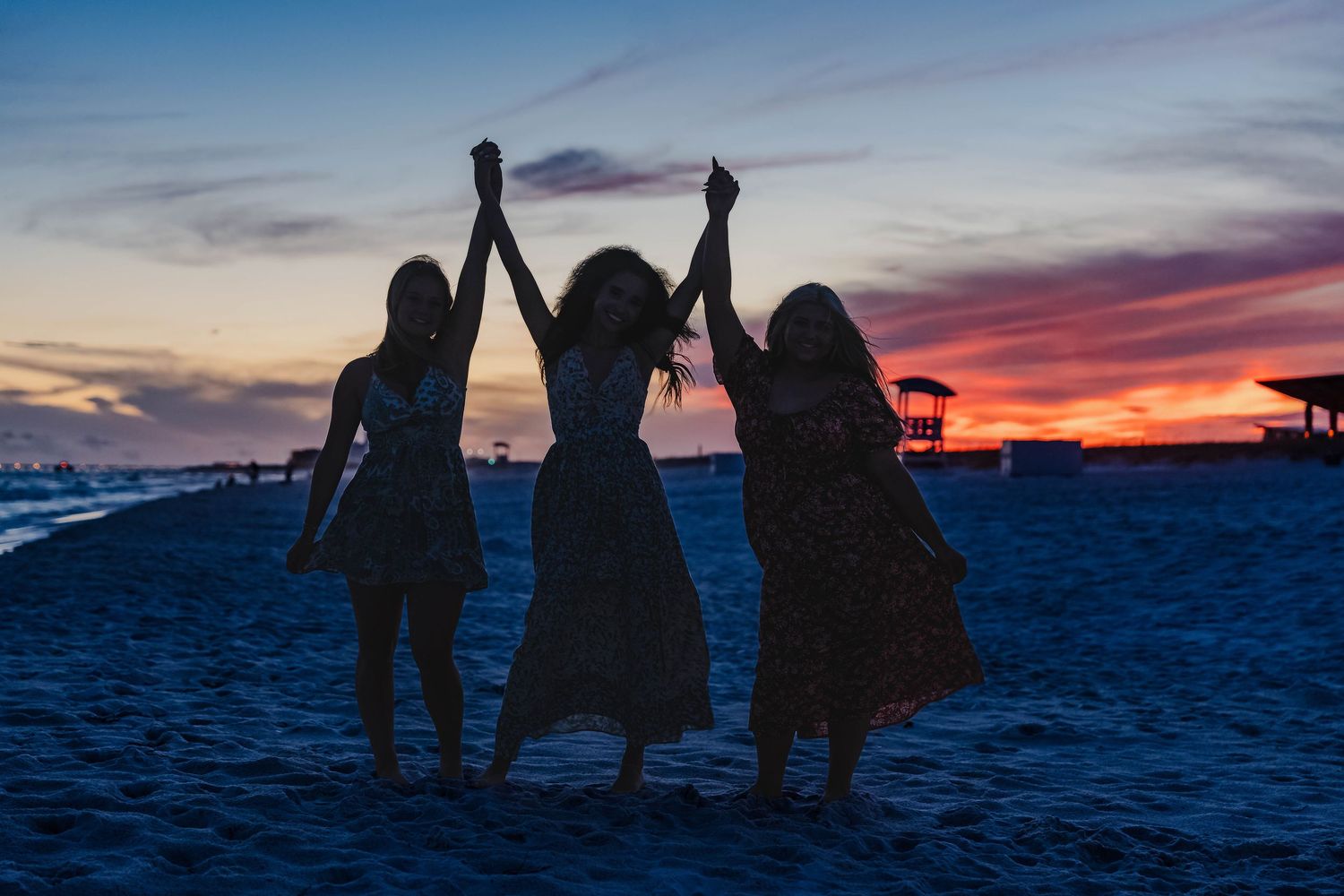 Three girls holding hands raised at sunset on a beach, silhouetted against a vibrant red and blue sky.