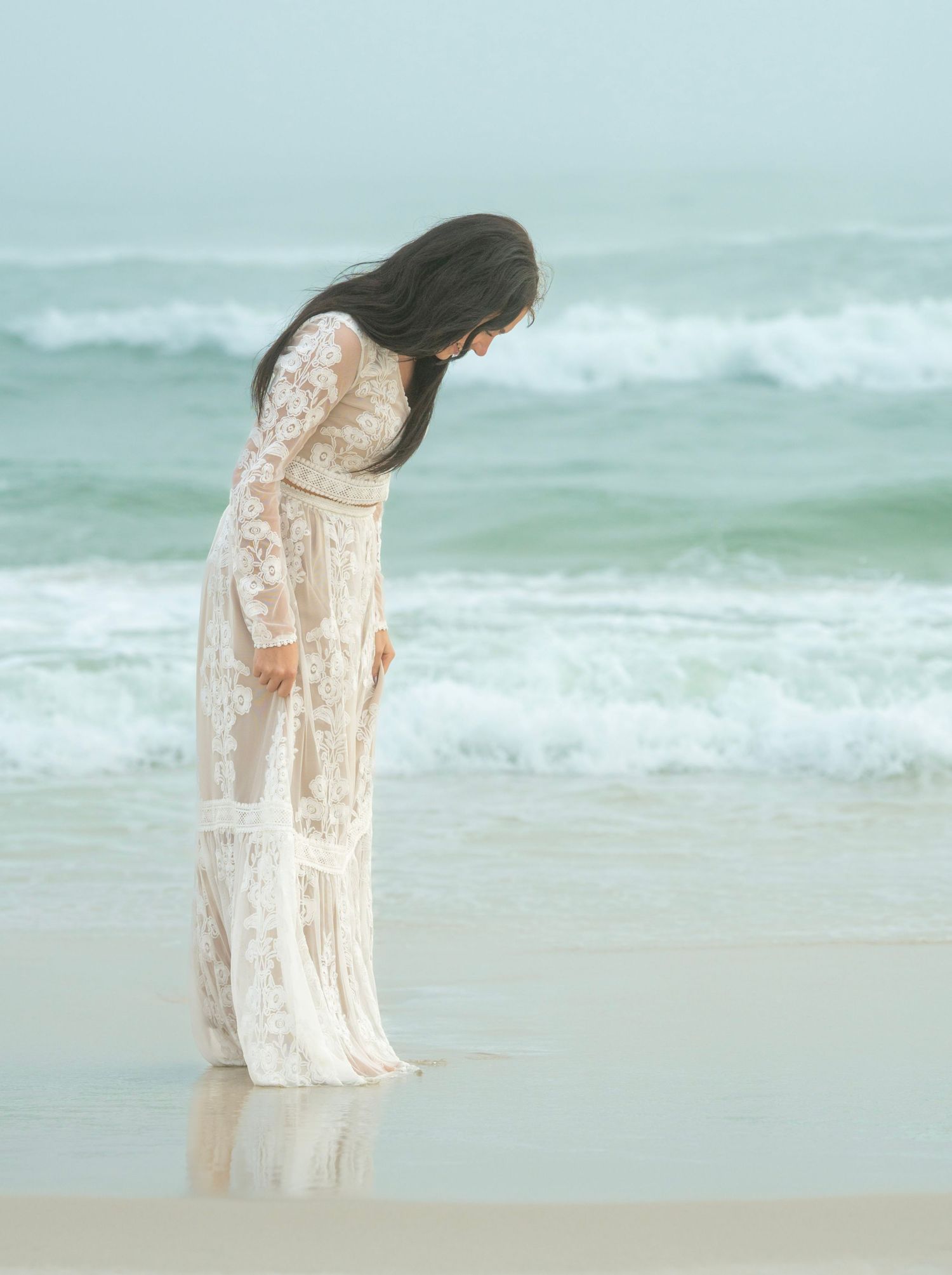 Person in white lace dress standing at ocean's edge looking down at waves.