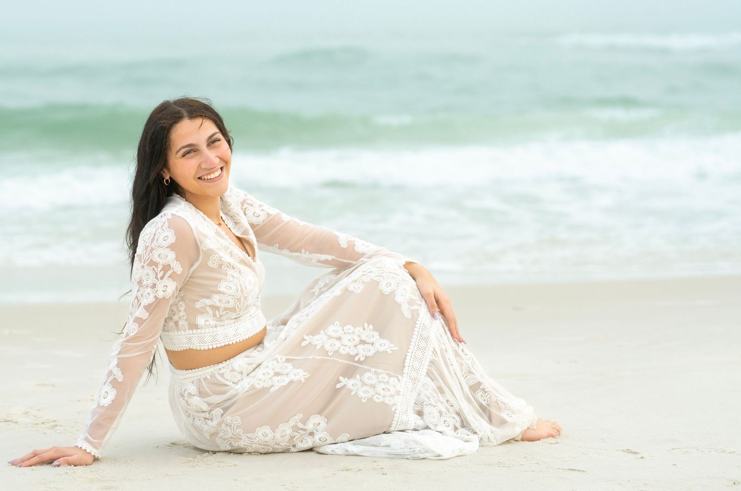 Woman in white lace dress poses gracefully on sandy beach with turquoise ocean waves in background.
