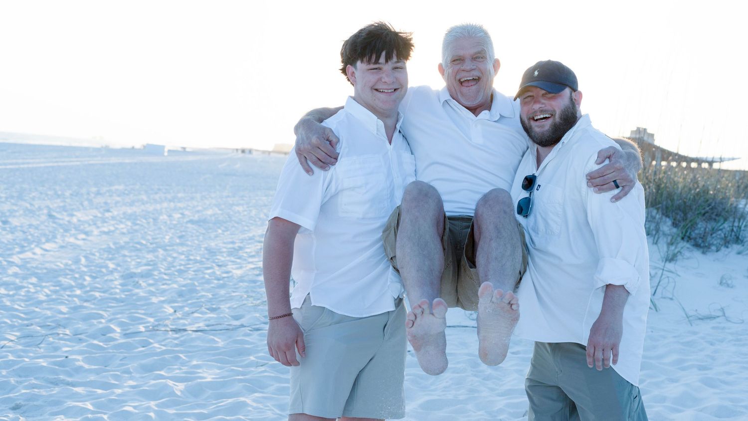 A group in white attire poses together on a snowy beach with palm trees and ocean waves in the background.