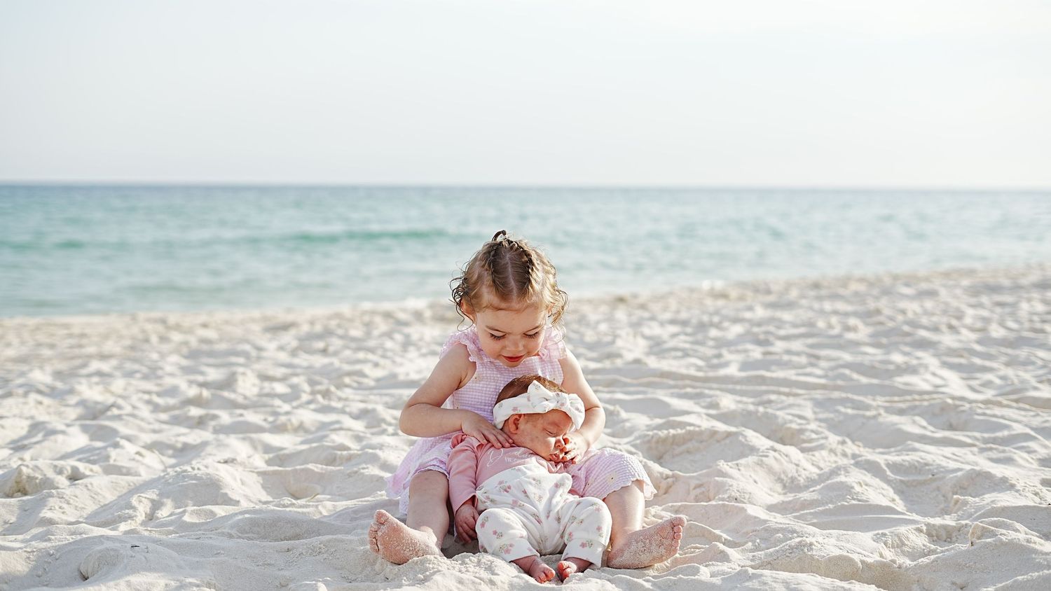 A small child sits on a pristine white sand beach near turquoise ocean waters while playing with a toy.