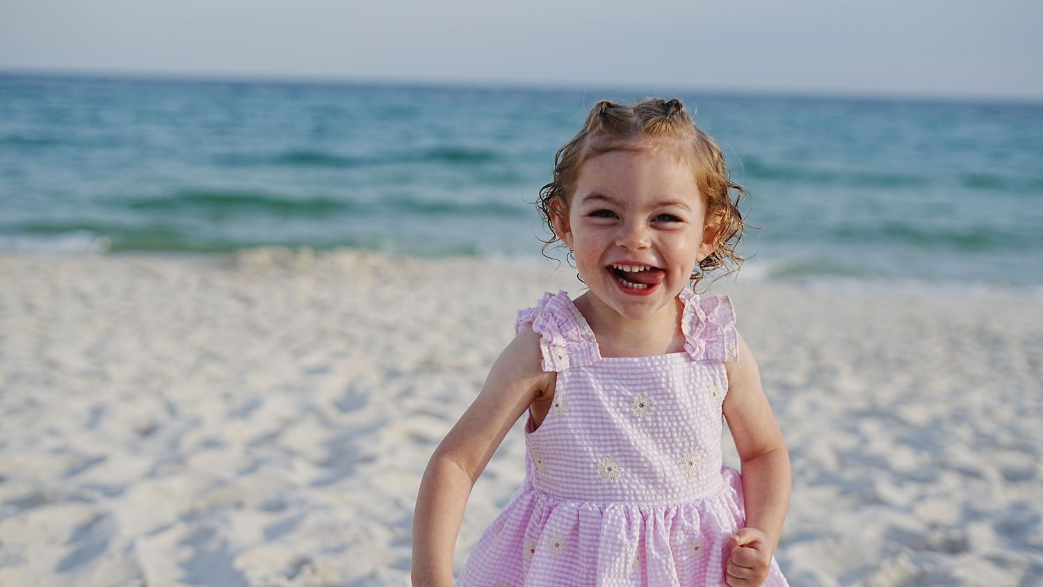 A child in a pink dress runs joyfully along a pristine white sand beach with turquoise ocean waters in the background.