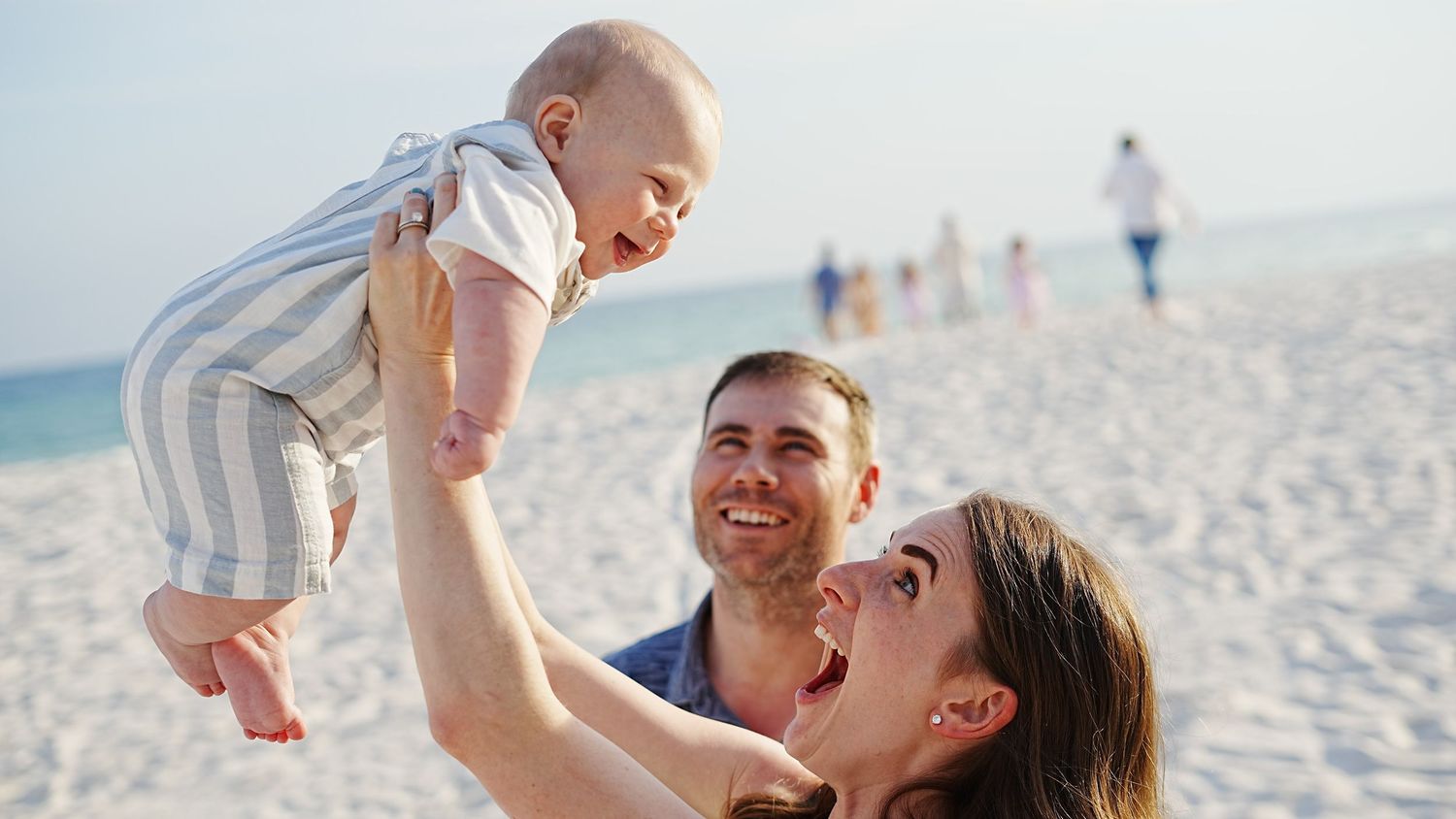 A joyful family moment at the beach with a baby being lifted up against a bright blue sky and ocean background.