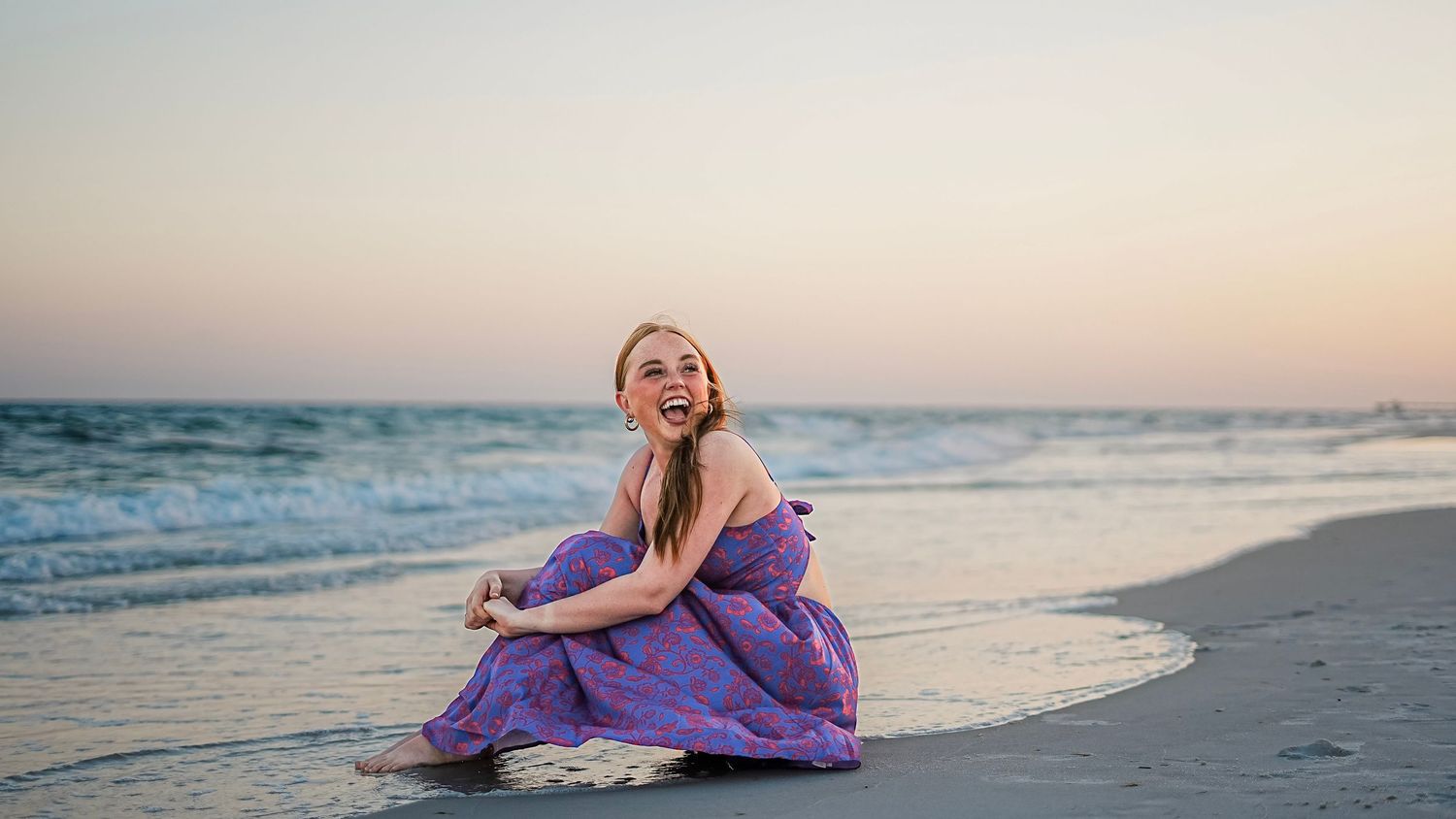 Woman in flowing purple dress sits gracefully on sandy beach at sunset with ocean waves in background.
