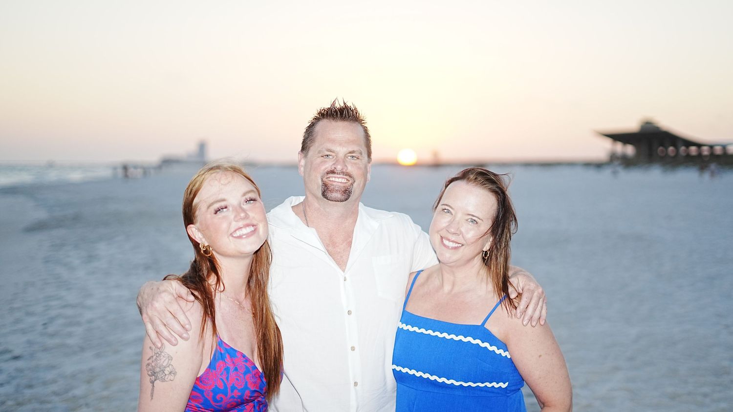 Three people laughing together on a beach at sunset with a pier visible in the background.