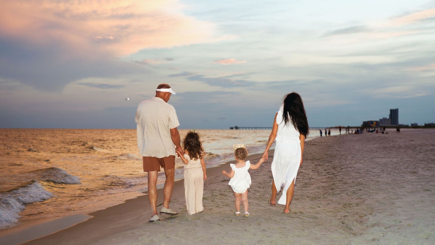 Family of four in white outfits walking hand-in-hand on a sandy beach at sunset with calm ocean waves.