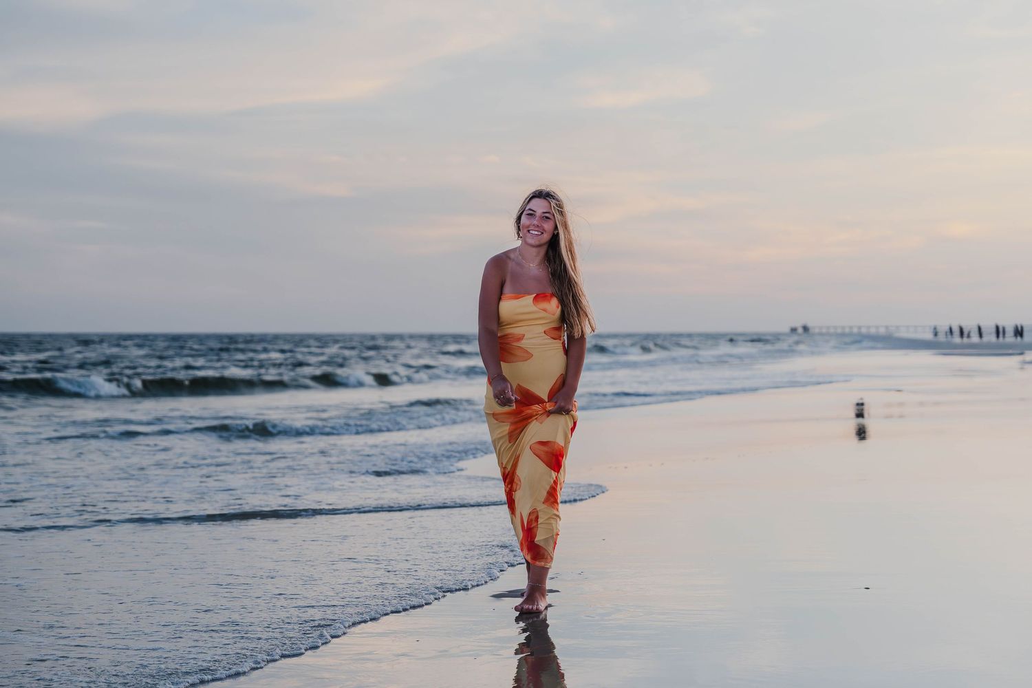 A young woman in a colorful strapless jumpsuit walks along a sandy beach at sunset with waves behind her.