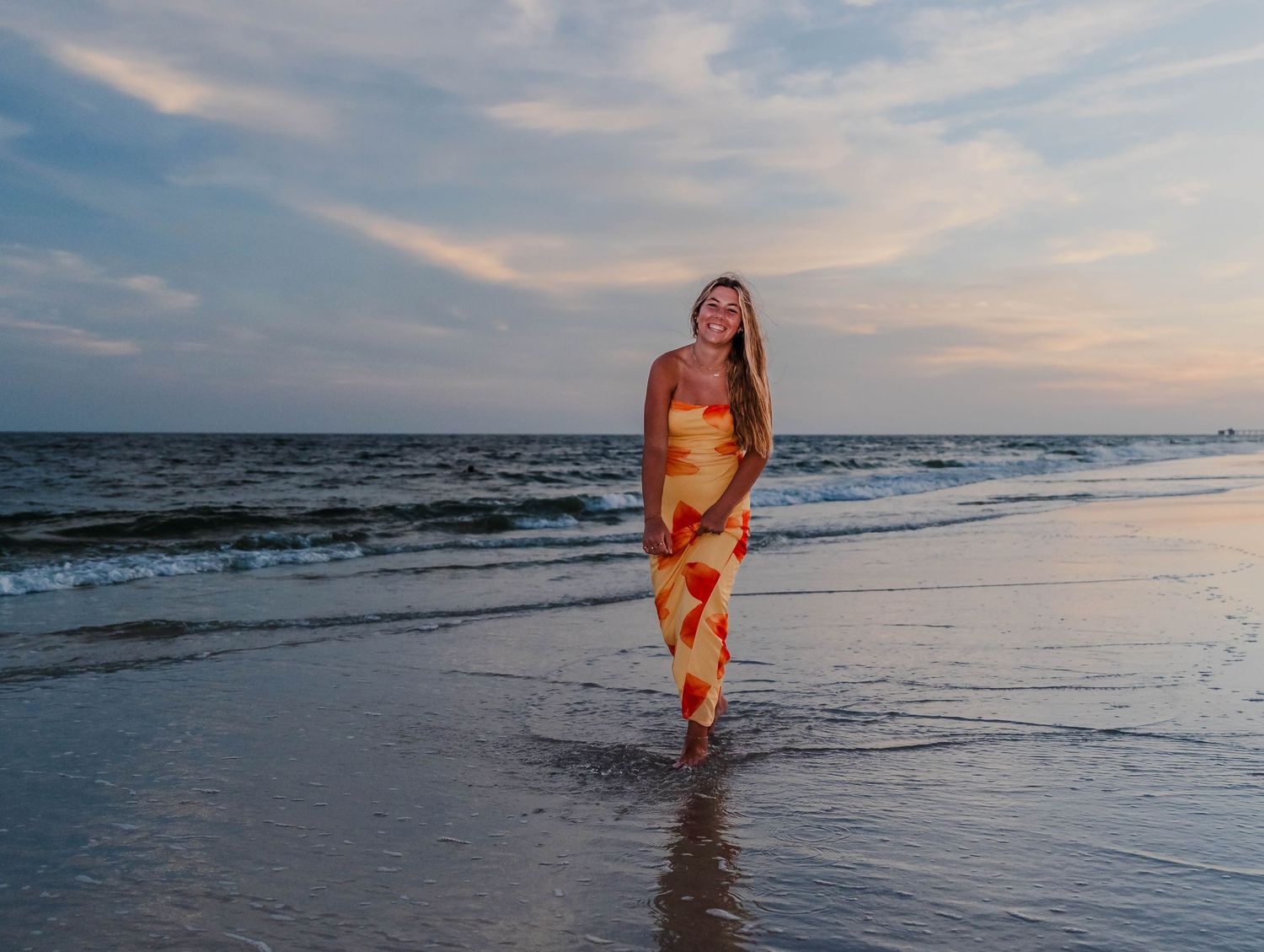Woman in colorful maxi dress walking on beach at sunset with ocean waves in background.