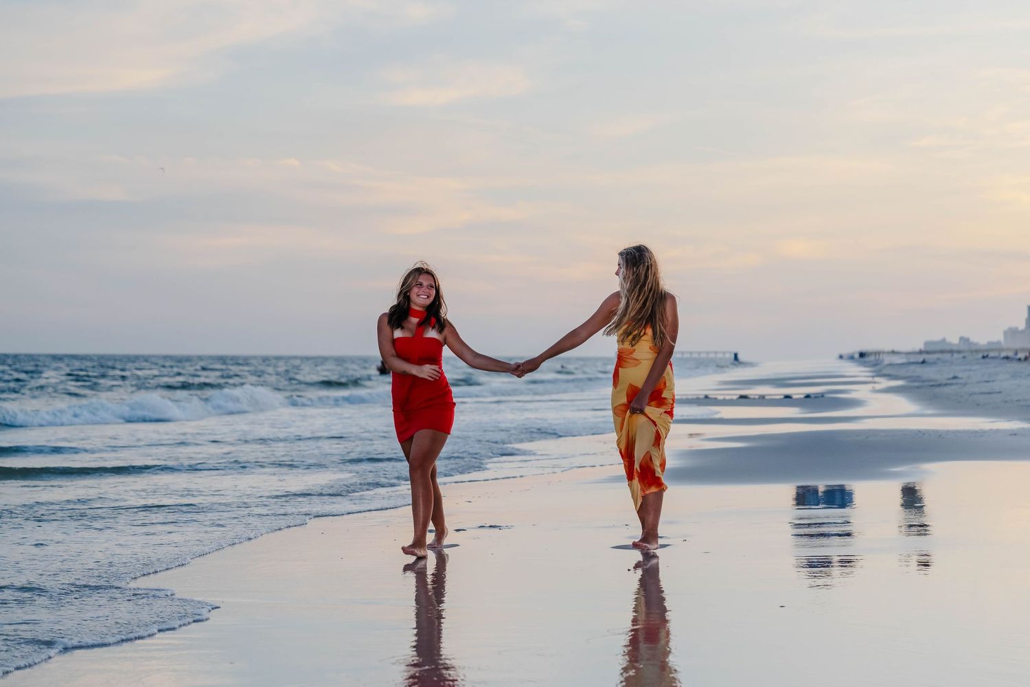 Two women in colorful dresses hold hands on a sandy beach at sunset with ocean waves in the background.