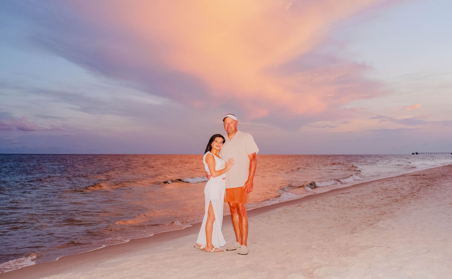 Couple in white outfits embracing on a sandy beach during a stunning pink and purple sunset over calm ocean waters.