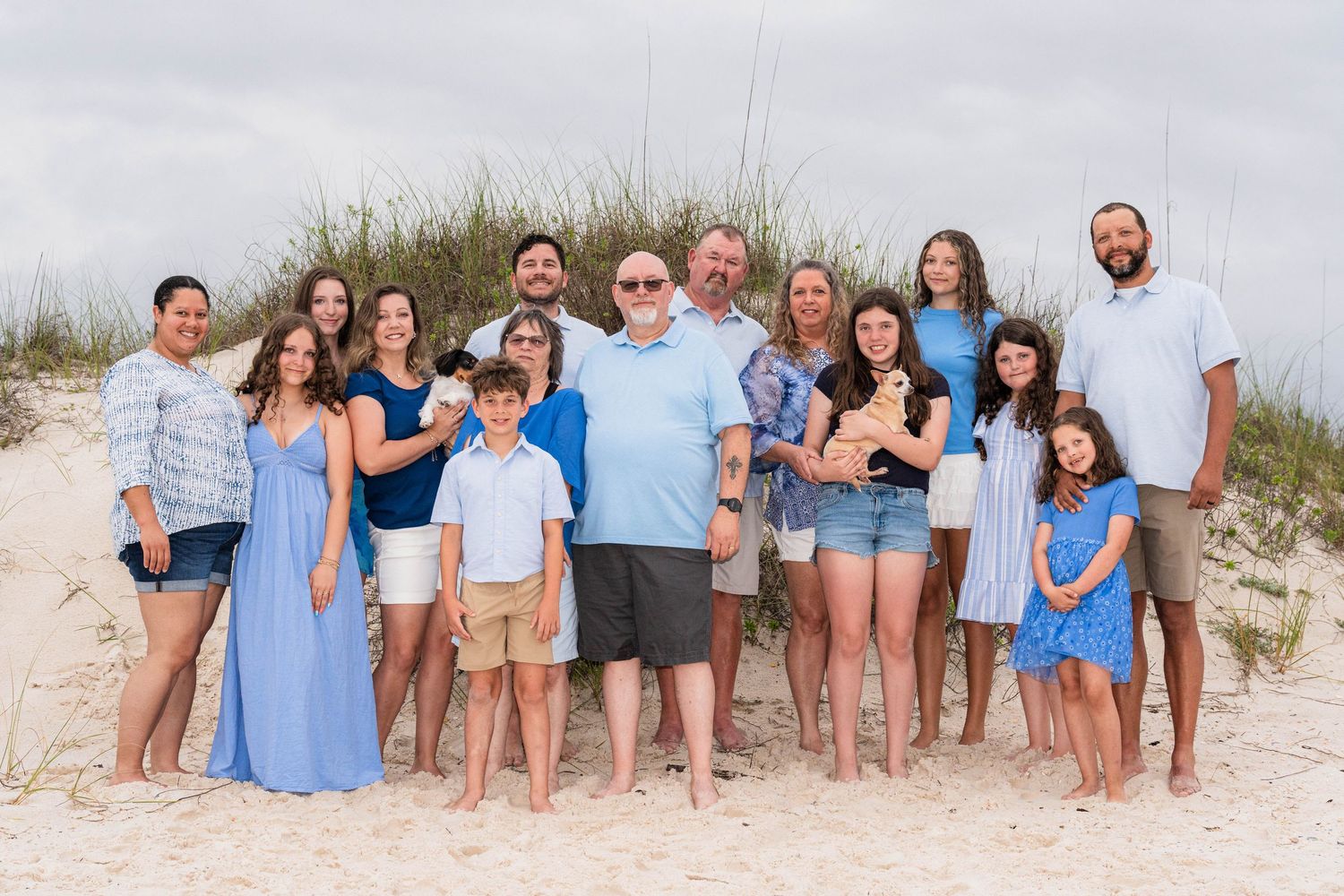 Large family group portrait on sandy beach with dunes, wearing coordinated blue and white outfits on overcast day.