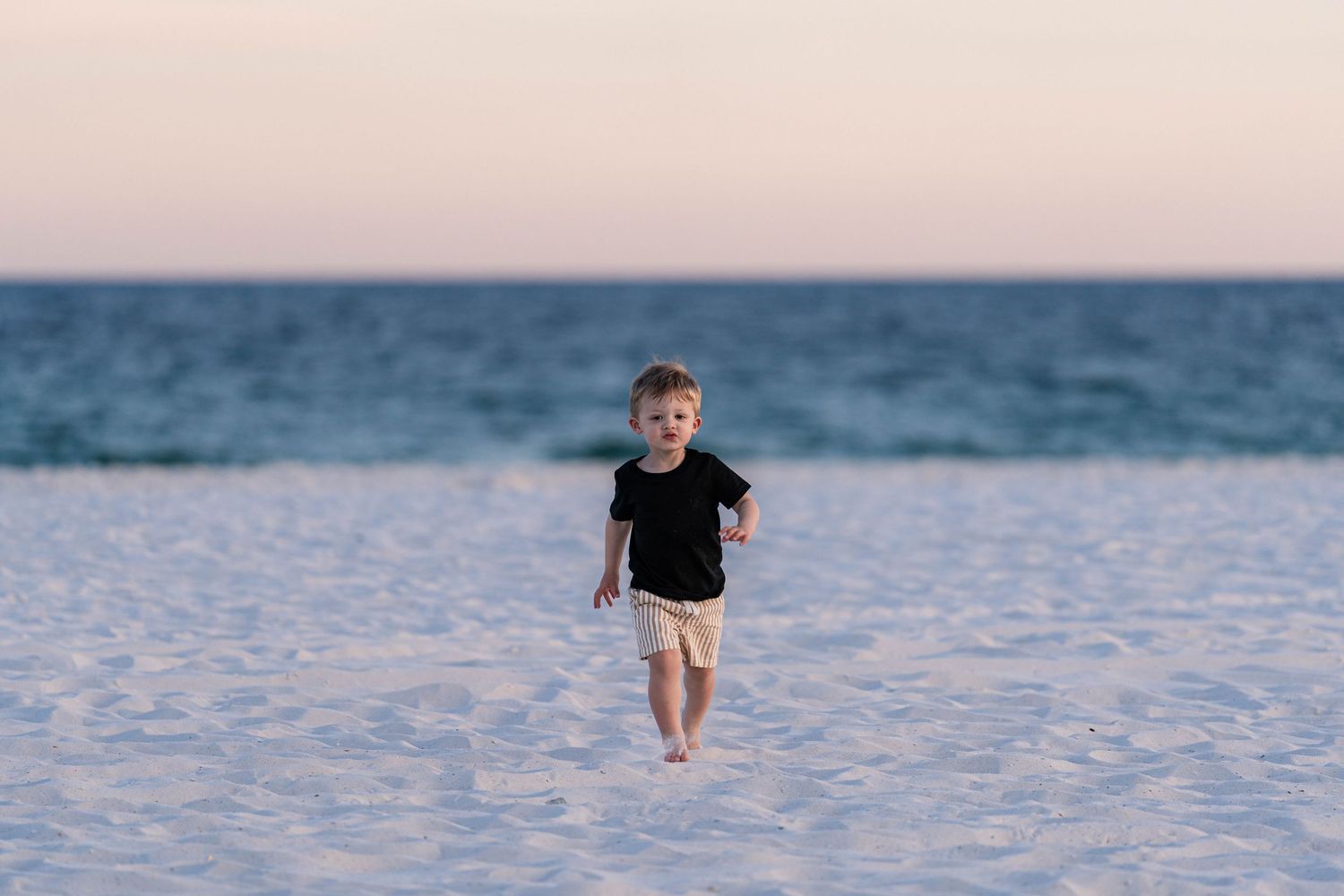 Toddler boy in black shirt and tan shorts running on white sand beach at sunset.