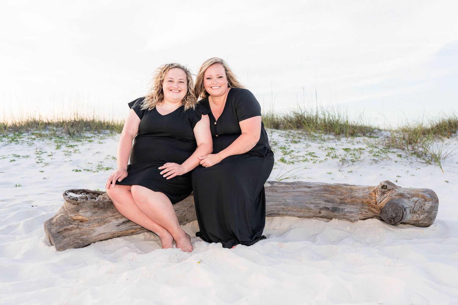 Two women in black dresses smile together on a driftwood log on a white sandy beach.