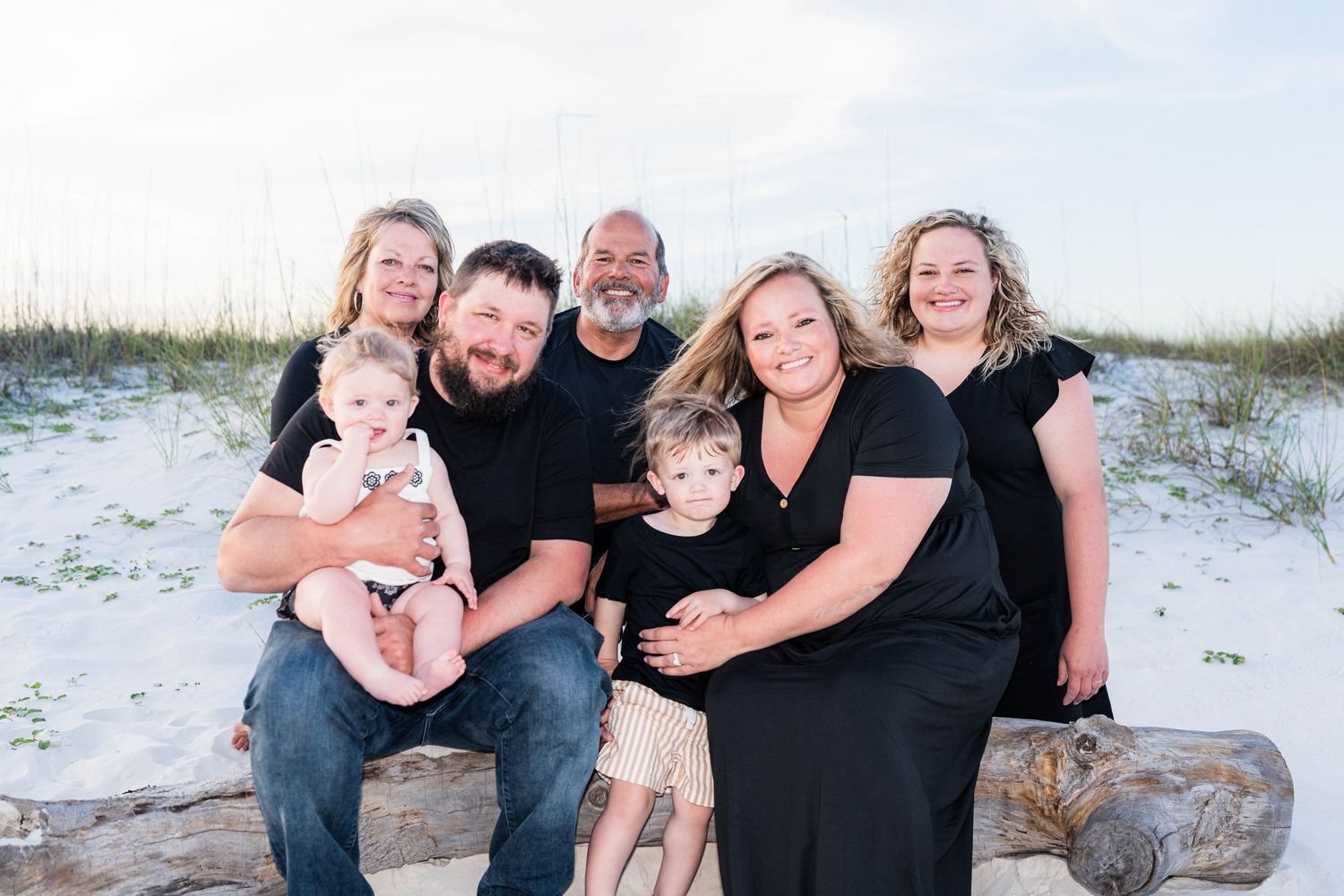 Family of six posing together on a sandy beach, smiling warmly during a professional outdoor photo session.