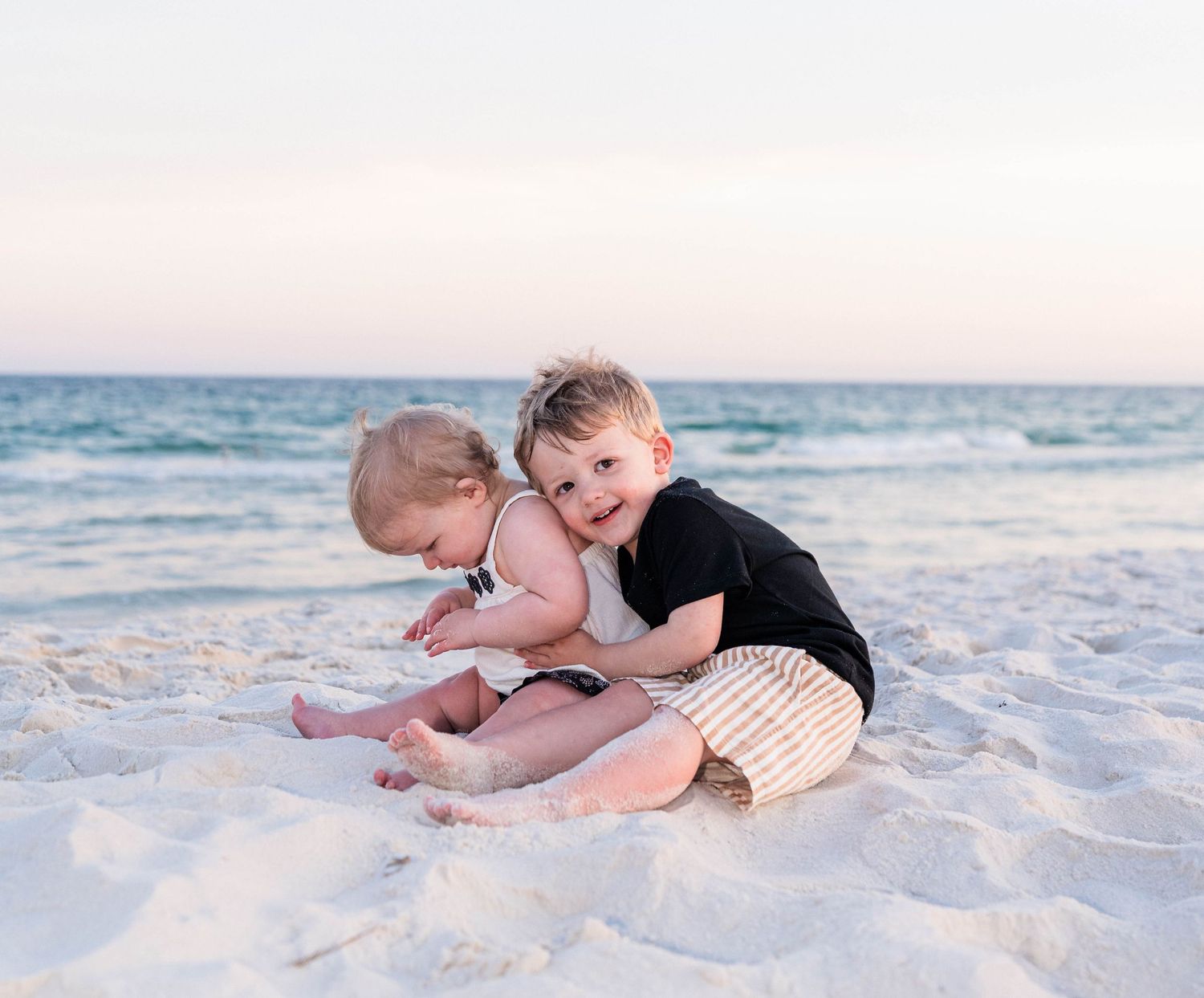 Two young siblings hugging and playing together on a white sandy beach at sunset.