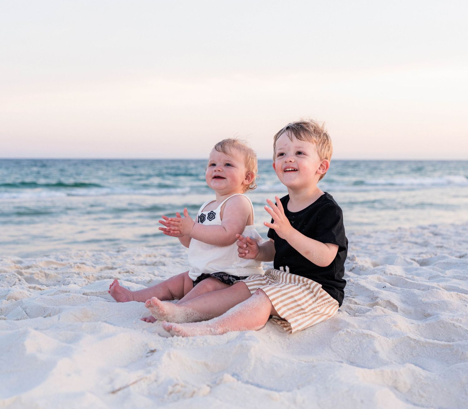 Two young children sitting together on white sand beach, smiling and clapping, with ocean waves in background.