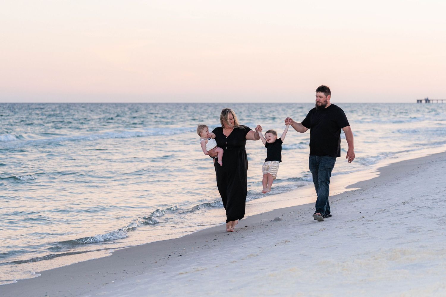 Family of four walking on a white sand beach at sunset, parents swinging two young children between them.