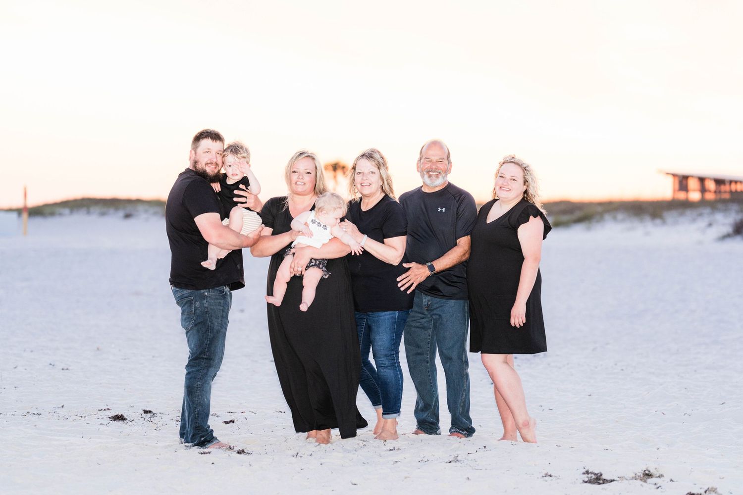 Family of six posing together on a sandy beach at sunset, wearing black and casual outfits, with two young children.