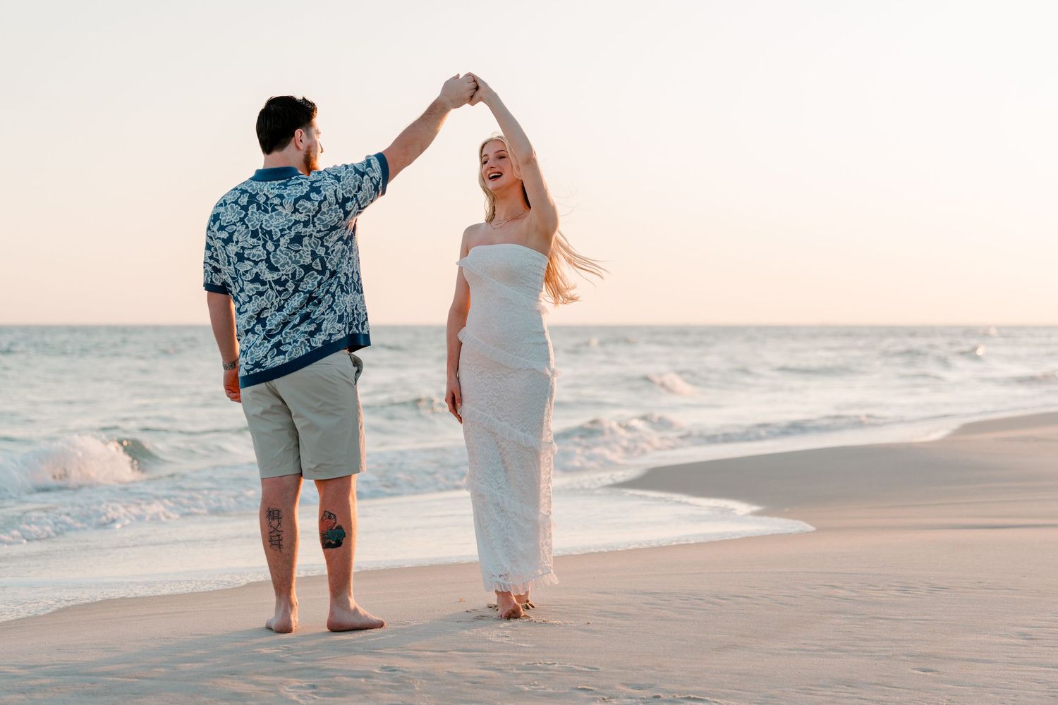 Couple dancing on beach at sunset, man spinning woman in elegant white dress by ocean waves.