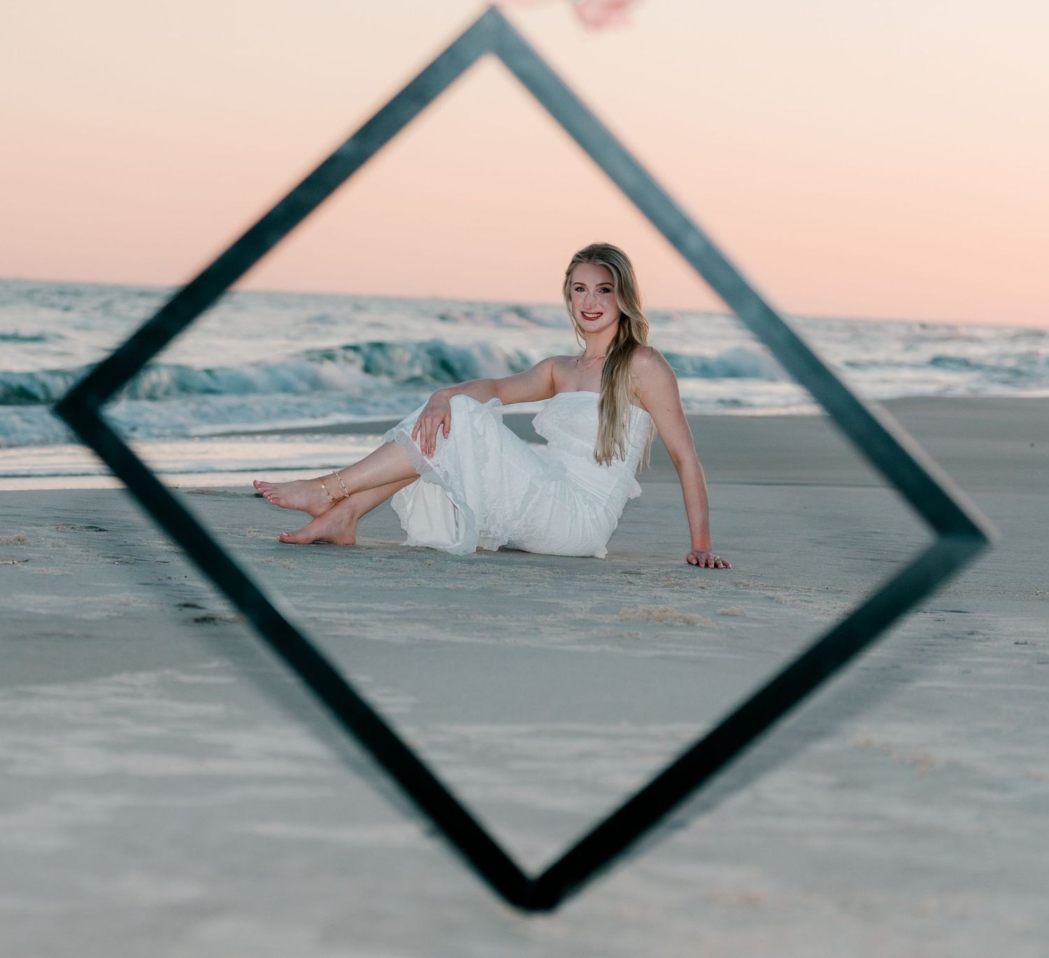 Woman in white dress sitting on sandy beach at sunset, framed by a diamond-shaped prop with ocean waves behind her.