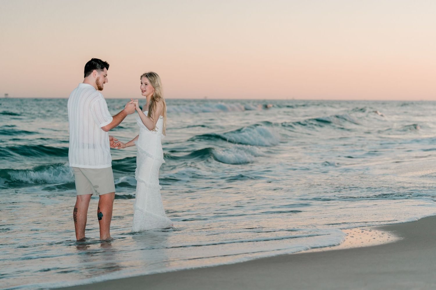 Couple standing in ocean waves at sunset, woman in white gown, man in white outfit.