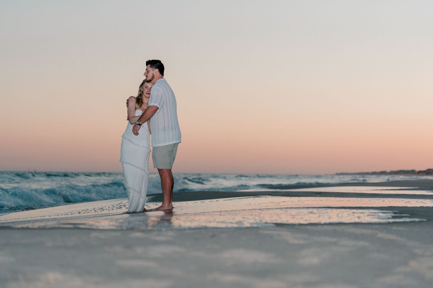 Couple embracing on a sandy beach at sunset with gentle ocean waves and a pastel pink sky backdrop.