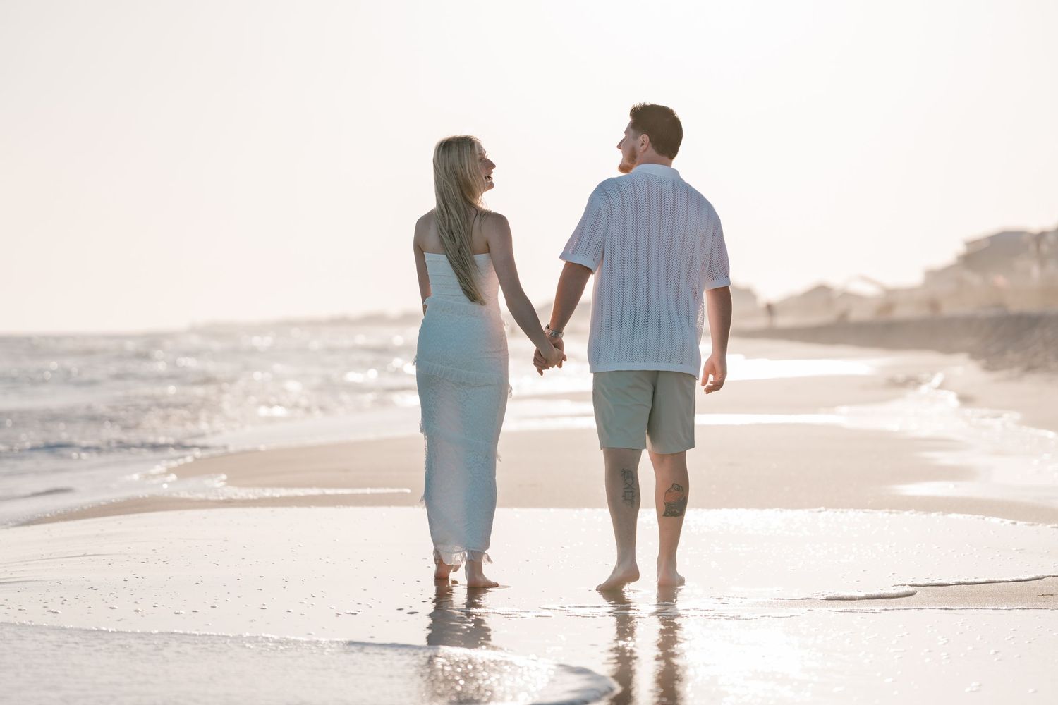 Couple holding hands walking along a sunny beach shore in matching light blue outfits.