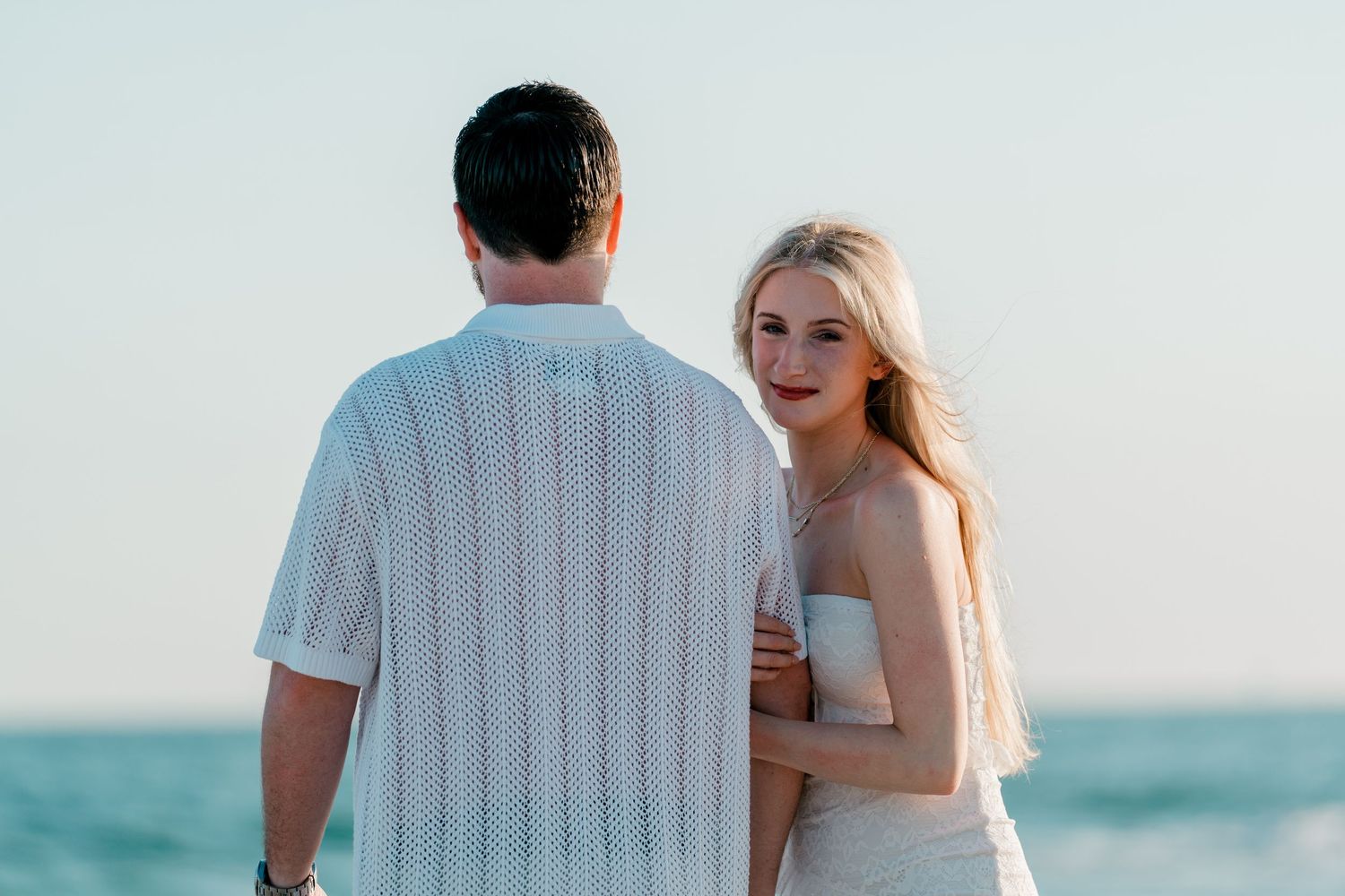 Couple standing on a beach at sunset, woman in white dress smiling over her shoulder at the camera.