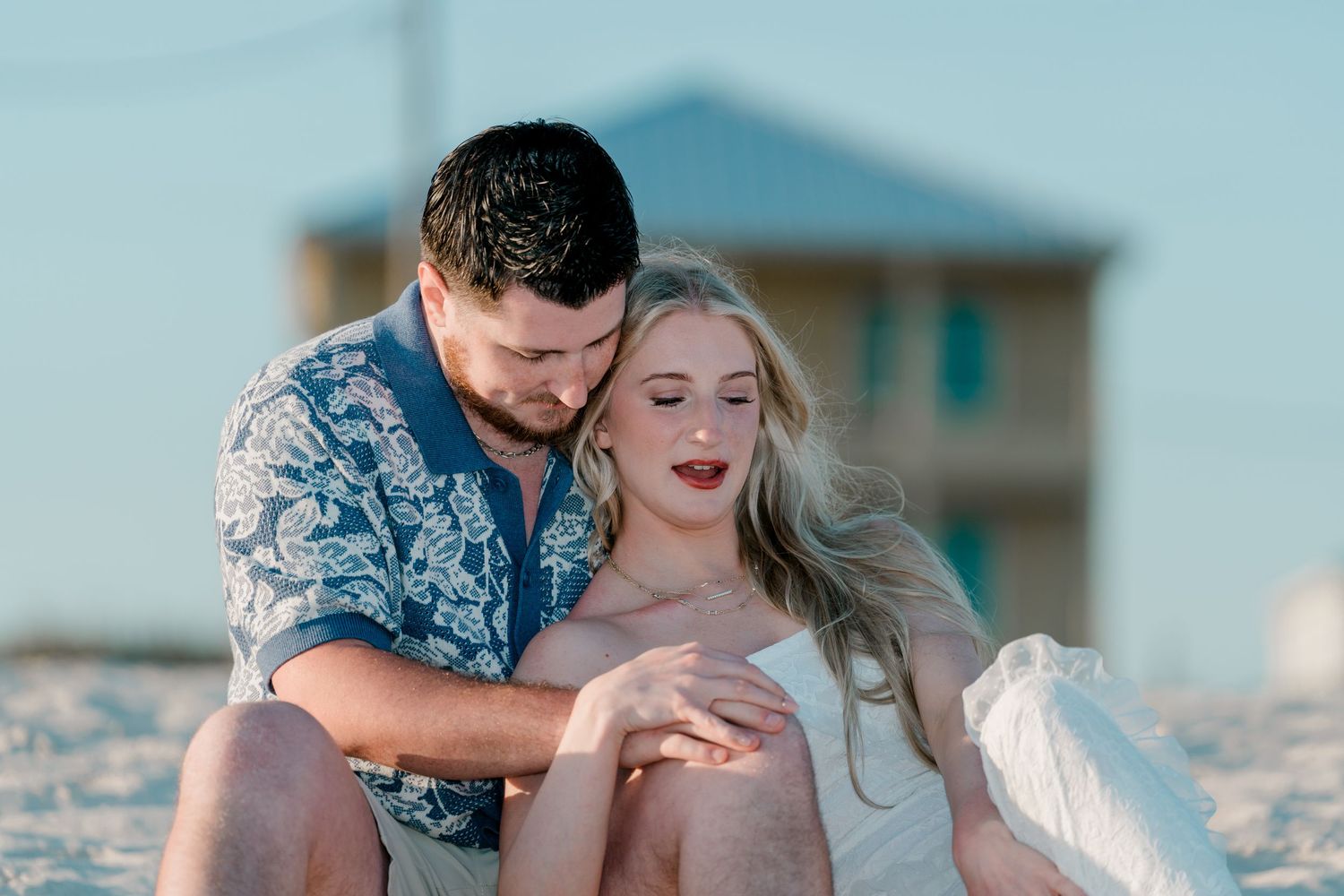 Couple sitting on beach sand at sunset, man in floral shirt embracing blonde woman in white dress.