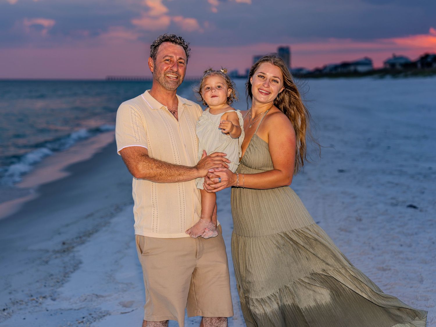 Family of three posing on a sandy beach at sunset, parents holding toddler with pink and purple sky backdrop.