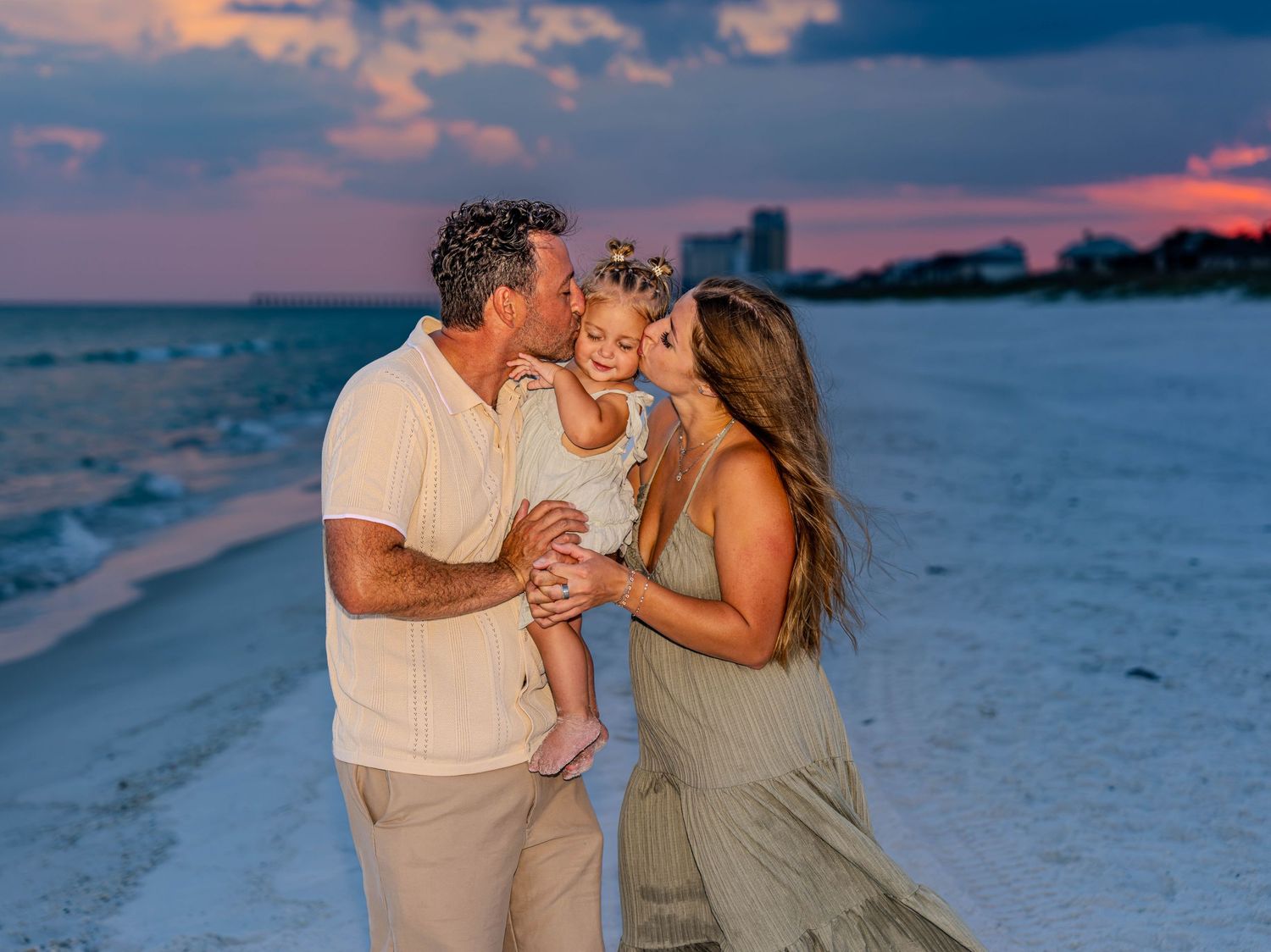 A family of three shares a tender moment on a white sand beach at sunset, parents kissing their toddler daughter.