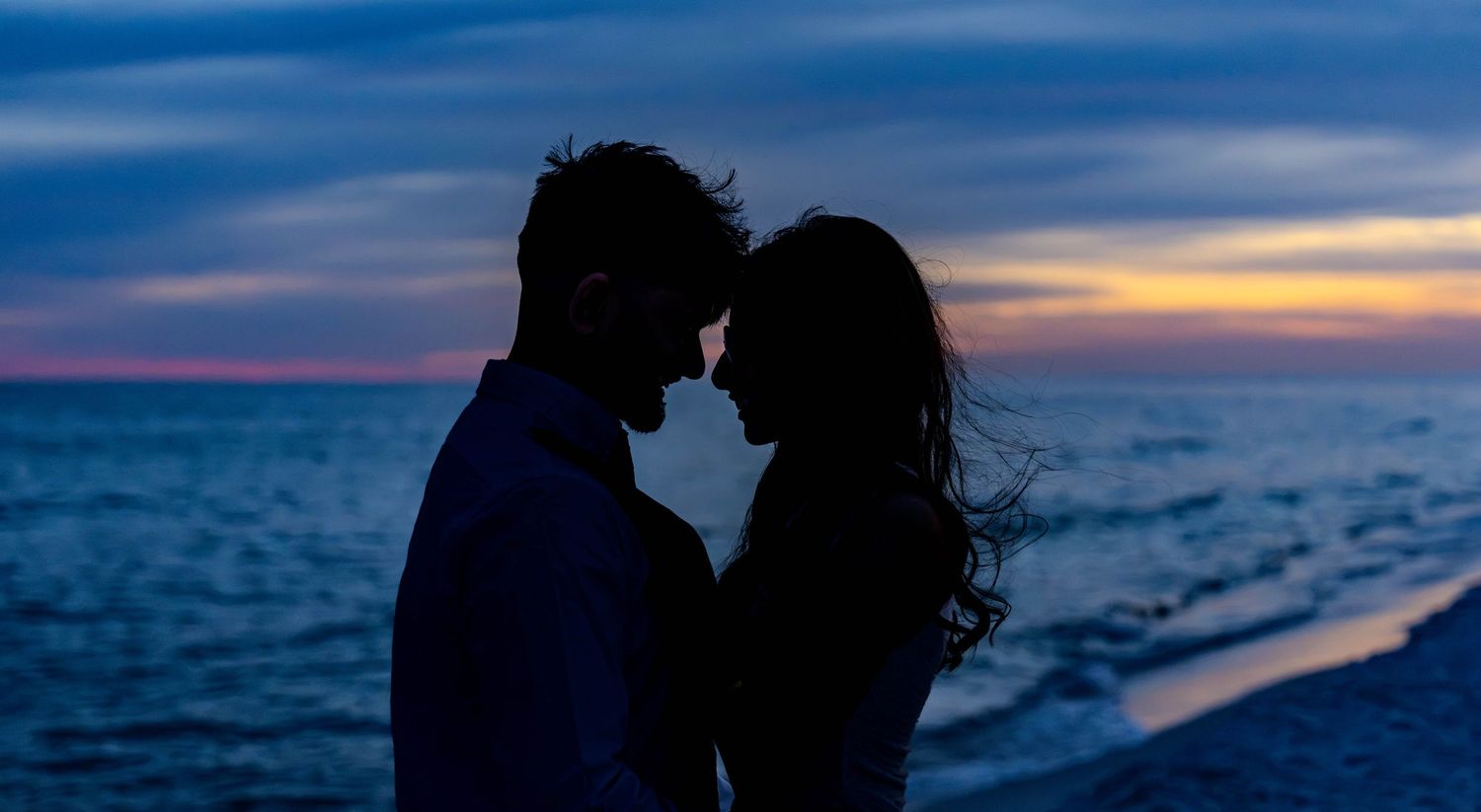 Romantic couple silhouette embracing on beach at sunset with colorful sky and ocean in background.