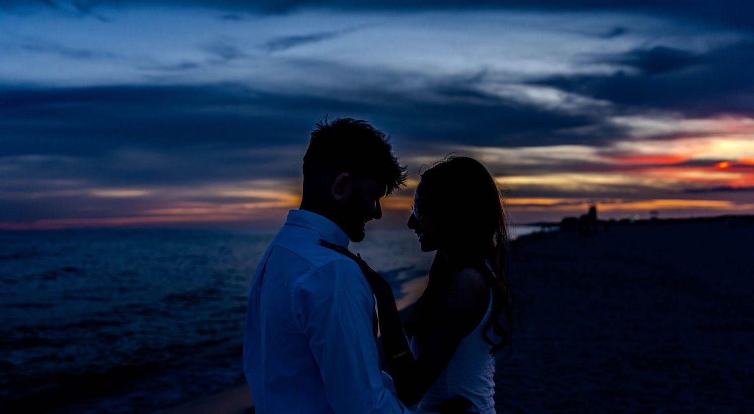 Romantic couple silhouetted against a vibrant sunset sky on a beach, sharing an intimate moment by the ocean.