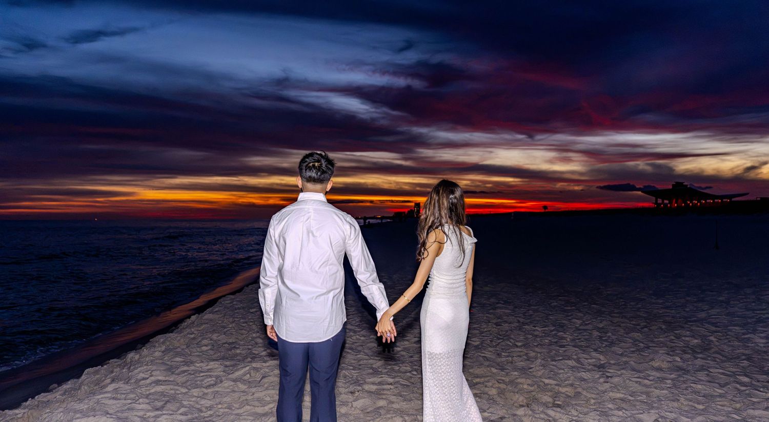 Couple holding hands on a sandy beach at sunset with vibrant red and purple sky over the ocean.