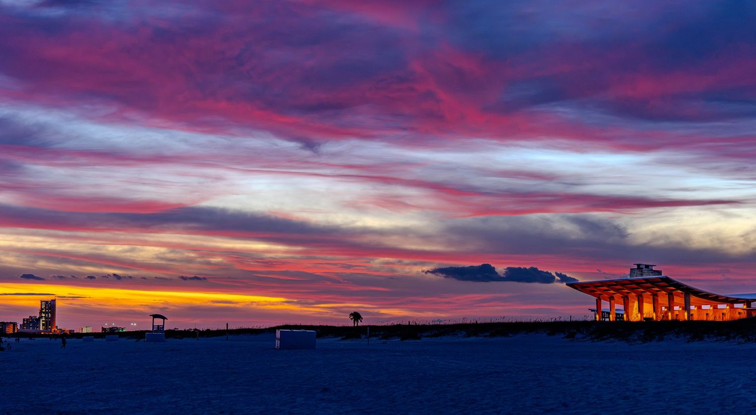 Vibrant sunset sky with pink and purple clouds over a coastal bridge and shoreline silhouette.