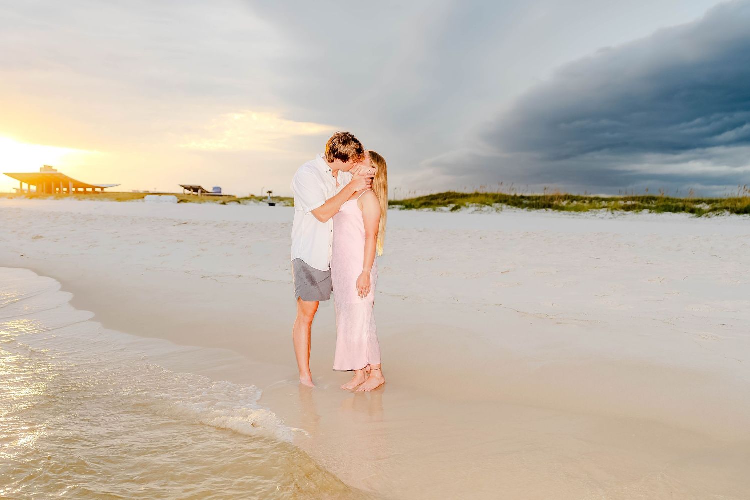 Couple embracing on white sand beach at sunset with dramatic storm clouds over calm ocean waters.