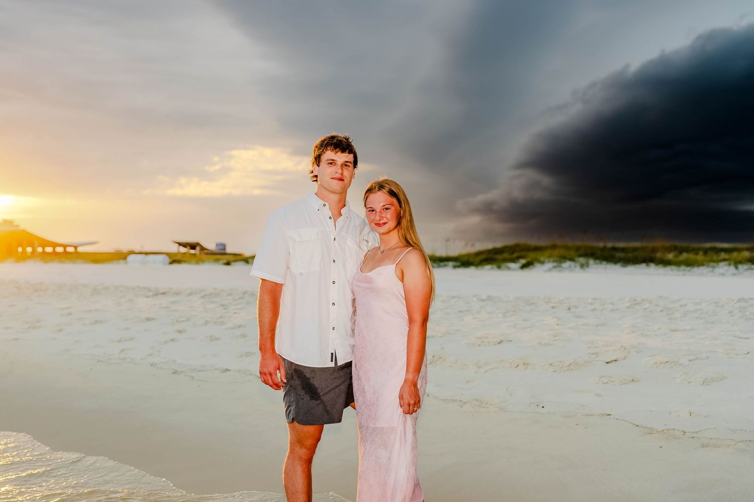 Couple smiling on white sand beach at sunset with dramatic storm clouds in background.