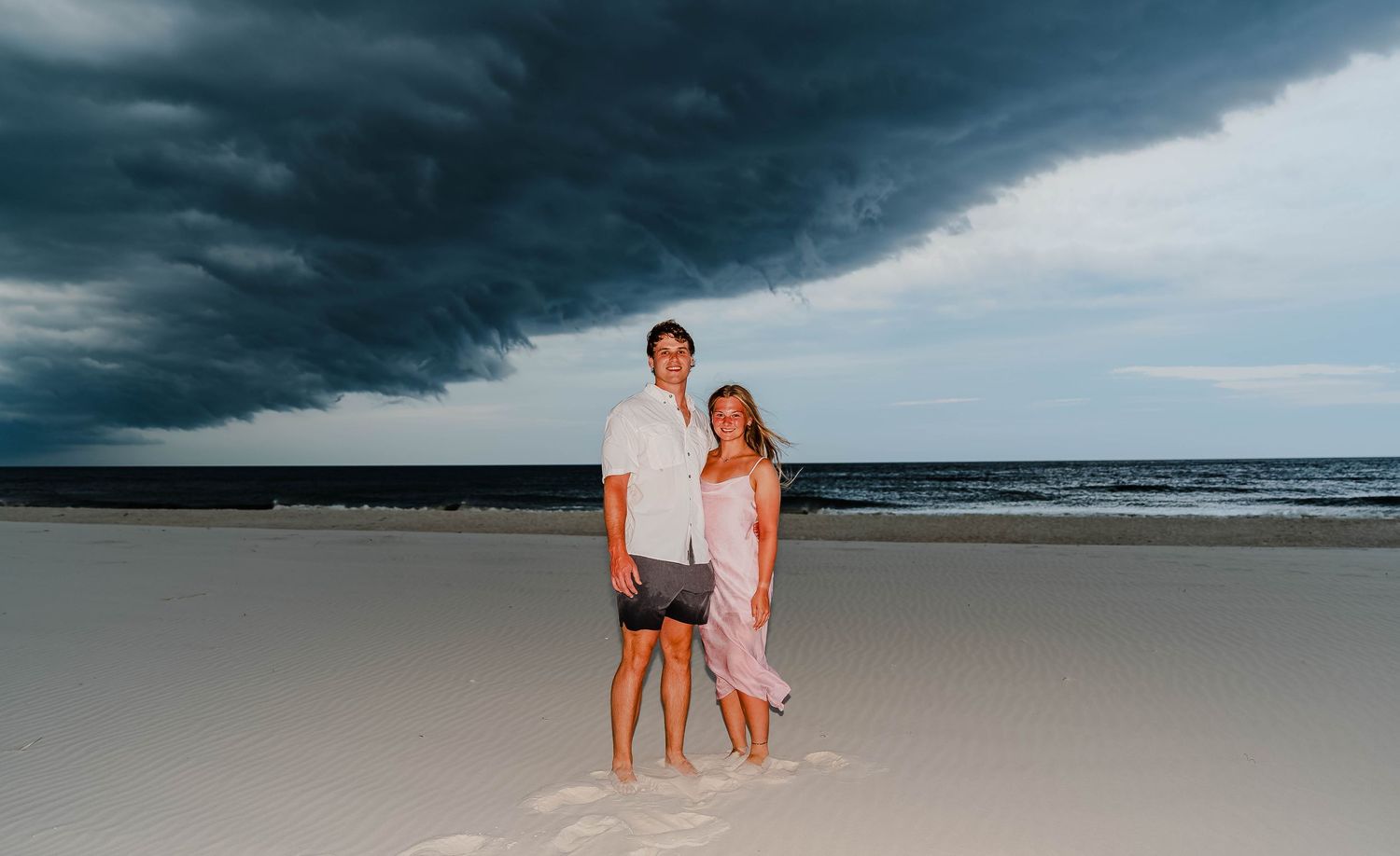 Couple posing on sandy beach with dramatic dark storm clouds over ocean behind them.