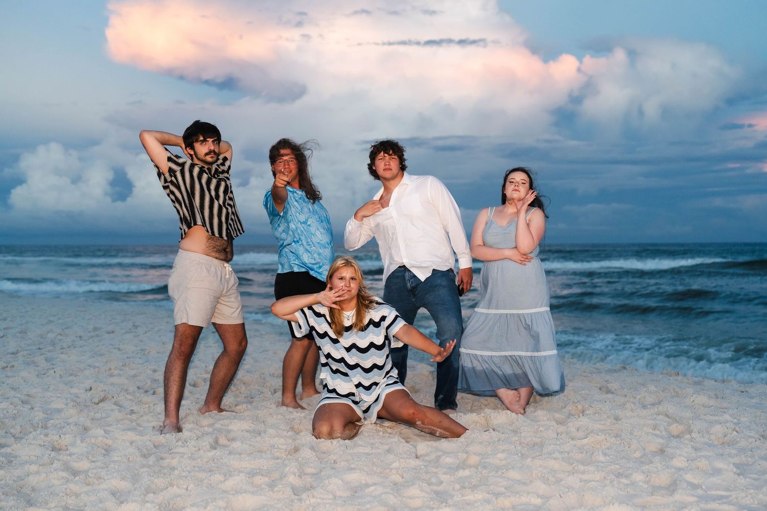 Five friends pose playfully on a sandy beach at sunset with dramatic cloudy skies and ocean waves behind them.