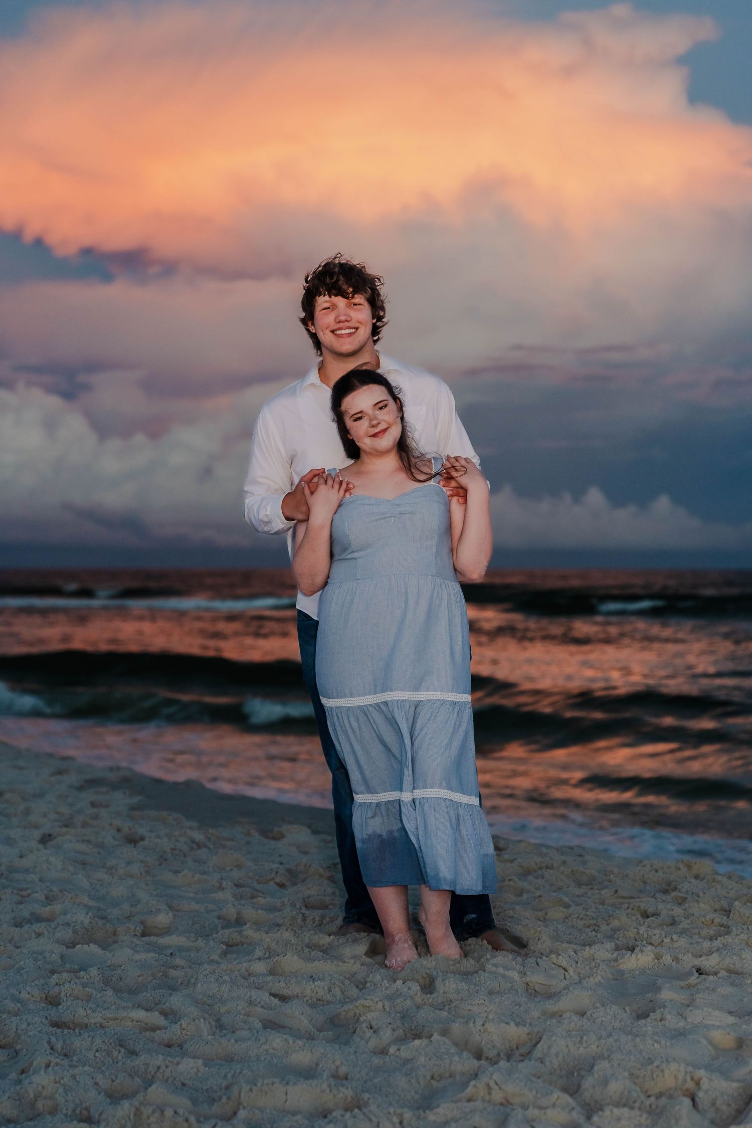 Couple embracing on sandy beach at sunset with dramatic pink and orange clouds over ocean waves.
