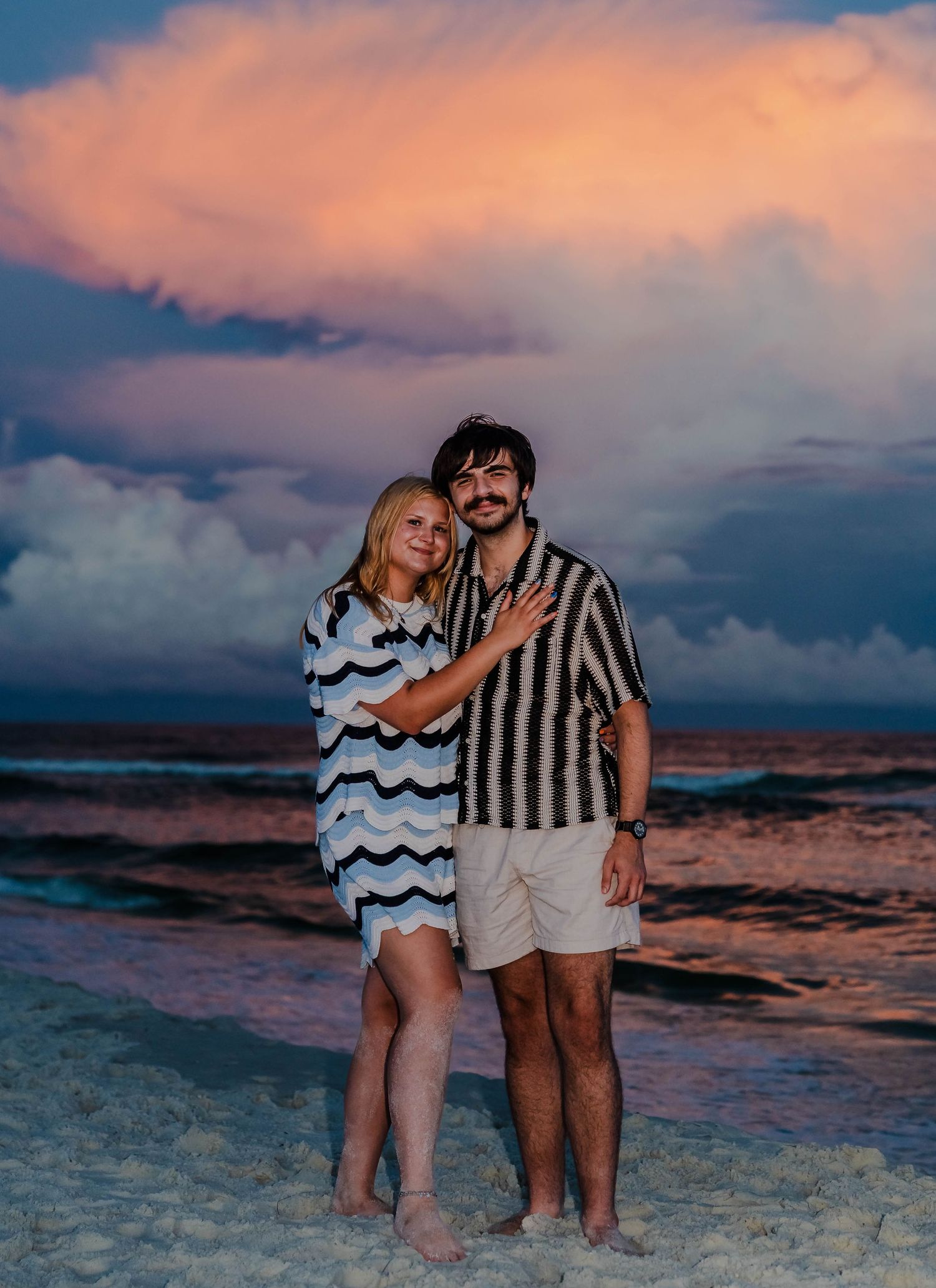 Couple embracing on a sandy beach at sunset with dramatic pink and blue cloudy sky over the ocean.