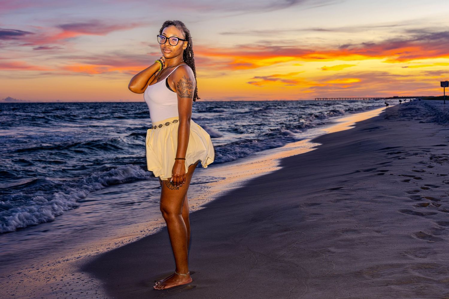 Woman in white dress standing on beach at sunset with tattoos and glasses, waves and colorful sky behind her.