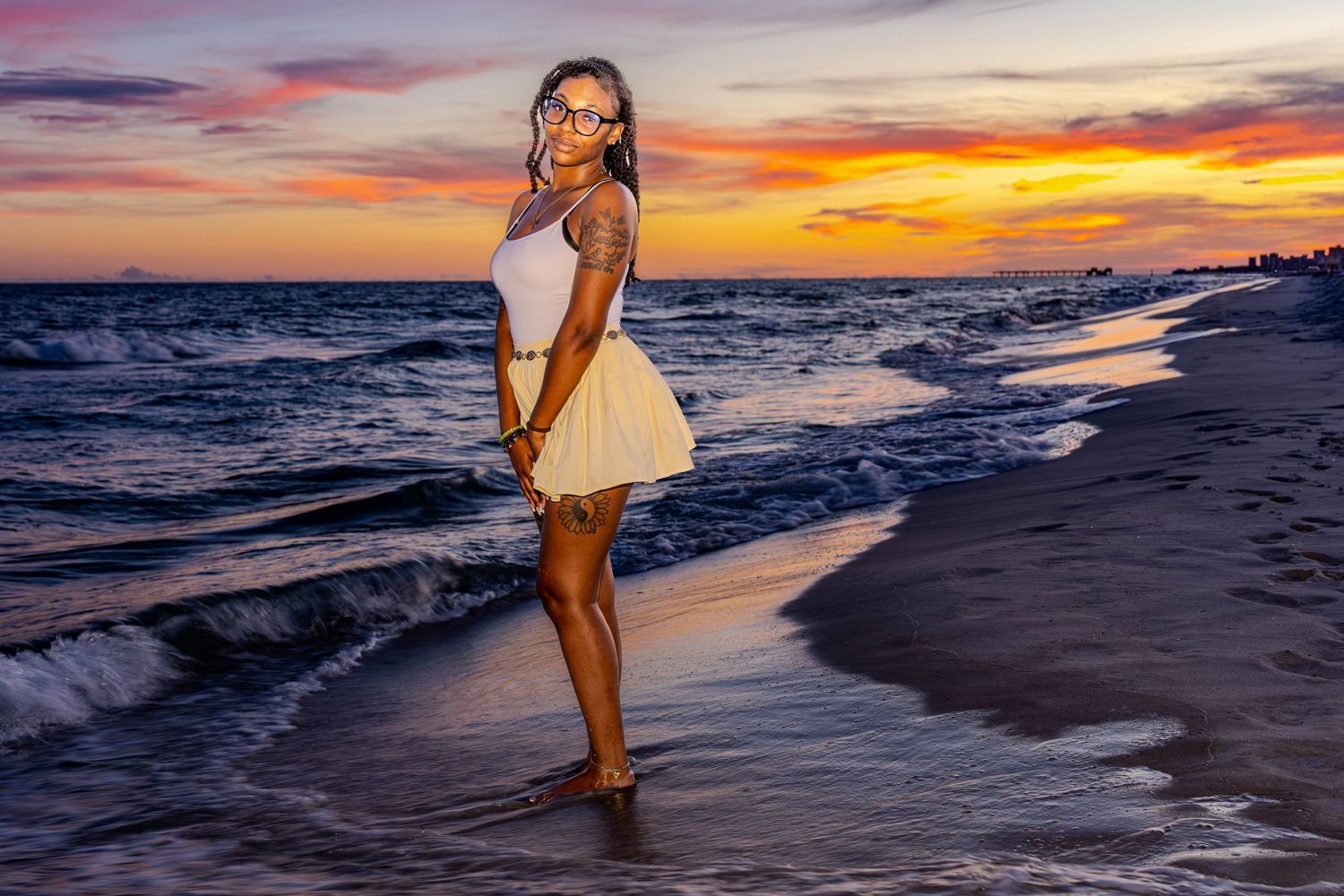 Woman in white dress standing on beach shore at stunning golden sunset with vibrant orange and purple sky.