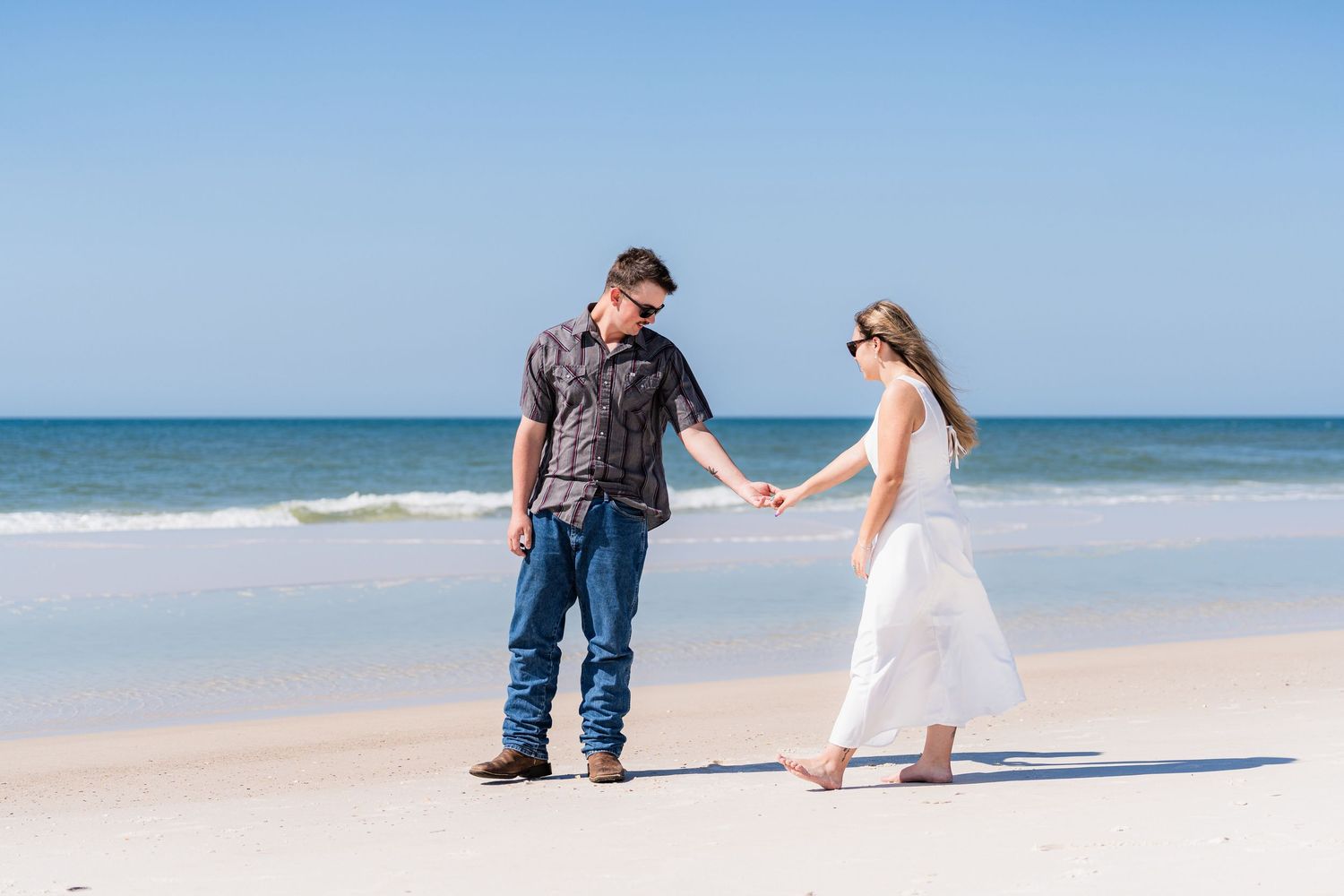 Couple holding hands on a white sandy beach with ocean waves, man in jeans and woman in white dress.